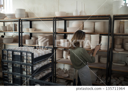 Female artisan in apron checking unglazed pottery at studio shelves with tablet, copy space Female artisan in apron checking unglazed pottery at studio shelves with tablet, copy space 135711964