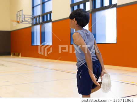 Asian boy stretching leg behind him holding shoe on gym court with basketball hoop, copy space 135711965