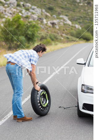 Middle-aged man lifting spare wheel beside sedan on mountain road using car jack, copy space Middle-aged man lifting spare wheel beside sedan on mountain road using car jack, copy space 135711968
