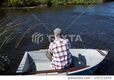 Senior angler wearing baseball cap sitting in small rowboat on river holding fishing rod over water Senior angler wearing baseball cap sitting in small rowboat on river holding fishing rod over water 135711970