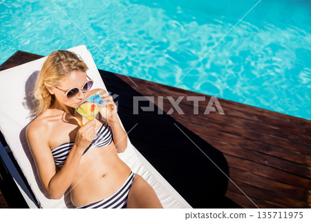 Woman in twenties relaxing on lounge chair beside turquoise pool holding cocktail glass, copy space 135711975