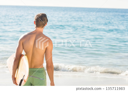 Shirtless man with green boardshorts holding longboard on shore gazing at turquoise sea, copy space 135711983