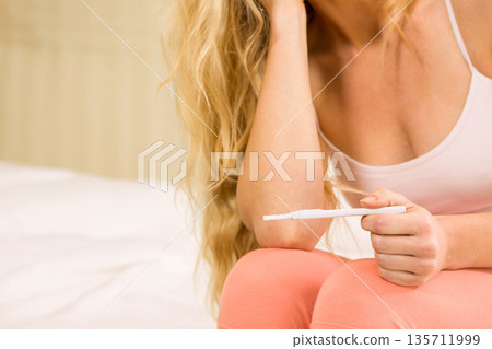 Blonde woman wearing tank top, leggings sitting on bed in softly lit bedroom holding pregnancy test Blonde woman wearing tank top, leggings sitting on bed in softly lit bedroom holding pregnancy test 135711999