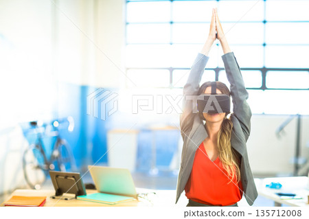 Female stretching arms upward wearing virtual reality headset at desk with laptop phone files bike 135712008
