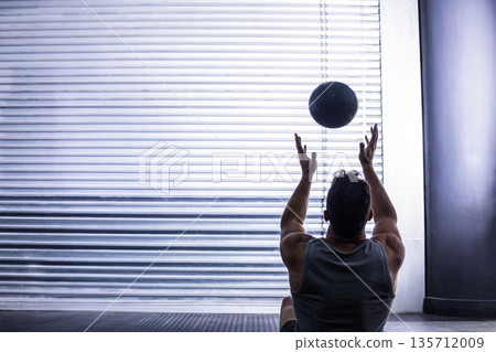 Mid adult man in sportswear sitting on gym floor raising medicine ball and slamming against shutter 135712009