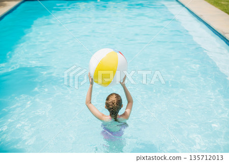 Female child holding inflatable beach ball while standing in backyard pool wearing purple swimsuit 135712013