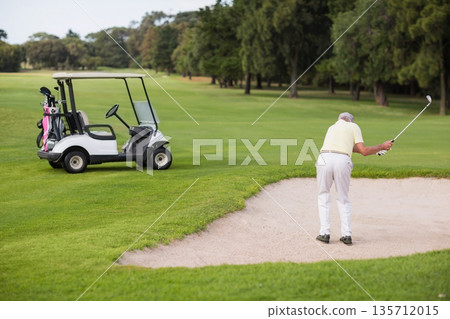 Senior man swinging golf club in sand bunker beside golf cart on golf course, copy space Senior man swinging golf club in sand bunker beside golf cart on golf course, copy space 135712015