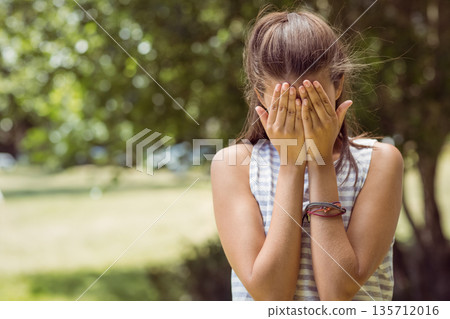 Adolescent female covering face with hands in park, wearing striped shirt, braided wristband 135712016
