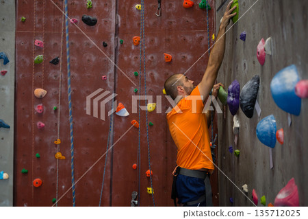 Male climber ascending gym wall gripping green hold on grey panel wearing blue harness, copy space Male climber ascending gym wall gripping green hold on grey panel wearing blue harness, copy space 135712025