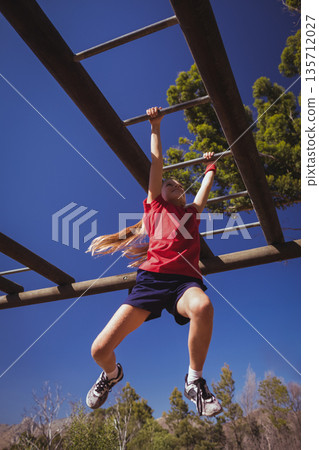 School-age girl hanging from wooden monkey bars at park under clear blue sky wearing red shirt School-age girl hanging from wooden monkey bars at park under clear blue sky wearing red shirt 135712027