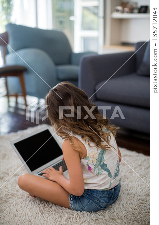 Female child wearing denim shorts sitting on rug in sunlit living room using laptop near sofa 135712043