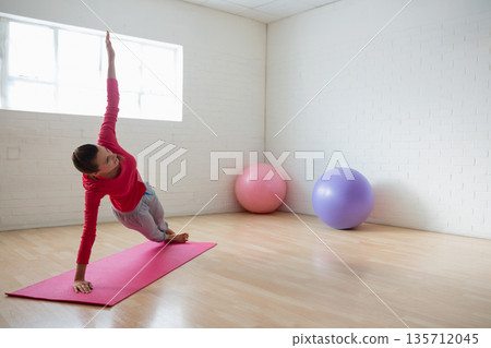 Woman holding side plank on pink yoga mat in studio with pink-purple exercise balls, copy space Woman holding side plank on pink yoga mat in studio with pink-purple exercise balls, copy space 135712045