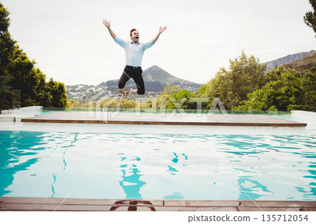 Man wearing blue shirt leaping off wooden deck beside turquoise pool raising arms toward peak 135712054