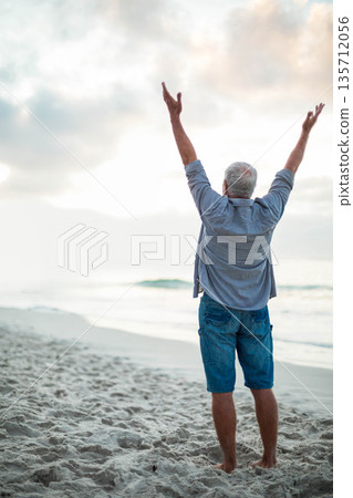 Senior man standing barefoot on sandy shoreline facing ocean raising arms toward sky with waves 135712056