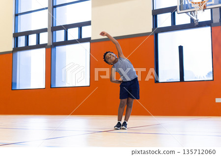 African American boy stretching in sportswear on gym court lines by windows and hoop, copy space 135712060