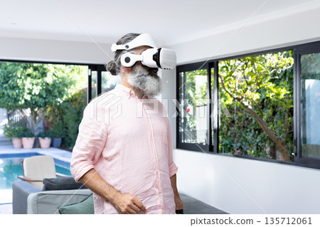 Senior man using virtual reality headset in bright living room with gray sofa, sliding glass doors Senior man using virtual reality headset in bright living room with gray sofa, sliding glass doors 135712061
