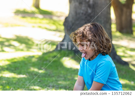 Curly-haired boy sticking out tongue while kneeling on grassy lawn in sunlit park, copy space Curly-haired boy sticking out tongue while kneeling on grassy lawn in sunlit park, copy space 135712062