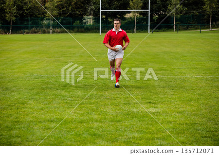 Male rugby player wearing red jersey sprinting while holding white ball on pitch before goalposts Male rugby player wearing red jersey sprinting while holding white ball on pitch before goalposts 135712071