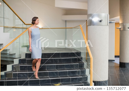 Adult woman descending black-tiled staircase with glass railing near support column in modern lobby Adult woman descending black-tiled staircase with glass railing near support column in modern lobby 135712077