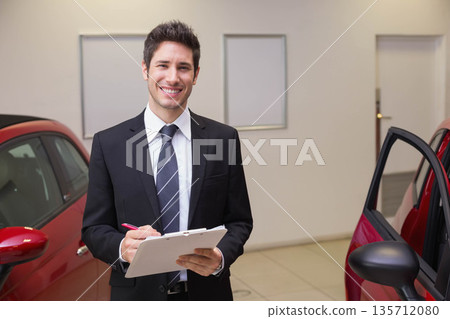 Adult male in suit standing between two red cars in dealership showroom holding clipboard and pen Adult male in suit standing between two red cars in dealership showroom holding clipboard and pen 135712080