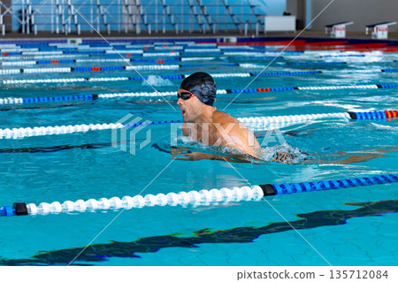 Male swimmer wearing cap and goggles gliding in breaststroke across lap pool toward starting blocks Male swimmer wearing cap and goggles gliding in breaststroke across lap pool toward starting blocks 135712084