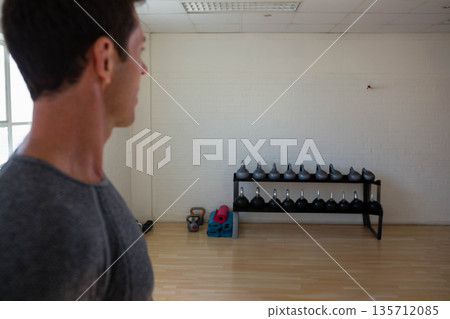 Man in grey t-shirt picking kettlebell from two-tier rack beside yoga mats in gym, copy space Man in grey t-shirt picking kettlebell from two-tier rack beside yoga mats in gym, copy space 135712085