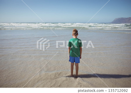 Boy standing knee-deep in waves wearing green shirt and blue shorts on shore beside distant cliff Boy standing knee-deep in waves wearing green shirt and blue shorts on shore beside distant cliff 135712092