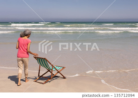 Senior African American woman positioning striped beach chair on wet sand at ocean edge, copy space Senior African American woman positioning striped beach chair on wet sand at ocean edge, copy space 135712094
