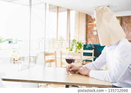 Man sitting at table in cafe holding wine glass with paper bag over head, copy space Man sitting at table in cafe holding wine glass with paper bag over head, copy space 135712098