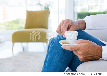 Hands are holding small white coffee cup with saucer and biscuit on sofa near large windows Hands are holding small white coffee cup with saucer and biscuit on sofa near large windows 135712099