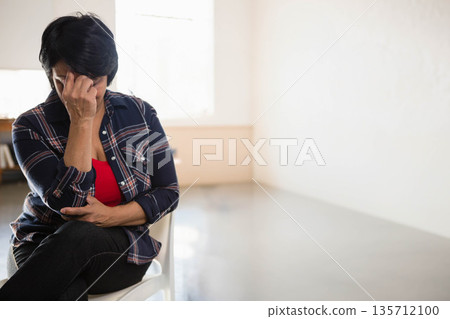 Indian mature woman pressing forehead sitting on white chair in bare room under window, copy space Indian mature woman pressing forehead sitting on white chair in bare room under window, copy space 135712100