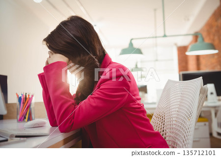 Asian woman in pink blazer leaning forward covering face amid colored pencils in office, copy space 135712101