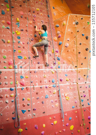 Female climber scaling climbing wall at climbing gym with colorful holds and harness, copy space Female climber scaling climbing wall at climbing gym with colorful holds and harness, copy space 135712105