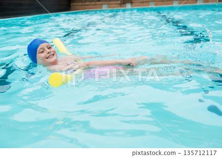 Female child floating on back using yellow noodle in outdoor pool sporting blue cap pink swimsuit 135712117