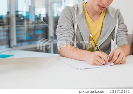 Man wearing grey hoodie writing in notebook with pencil in office with glass partitions, copy space 135712121