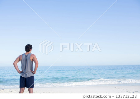Man wearing sportswear standing on sandy shoreline facing ocean under clear sky, copy space Man wearing sportswear standing on sandy shoreline facing ocean under clear sky, copy space 135712128