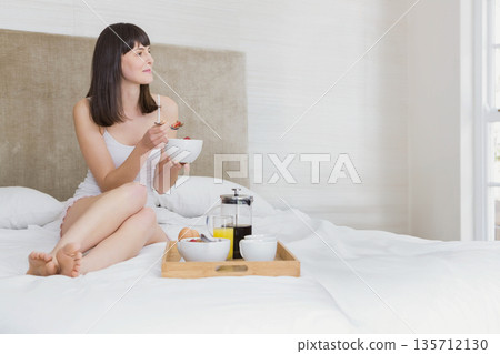 Woman sitting on bed at home holding bowl beside breakfast tray with coffee press, copy space 135712130