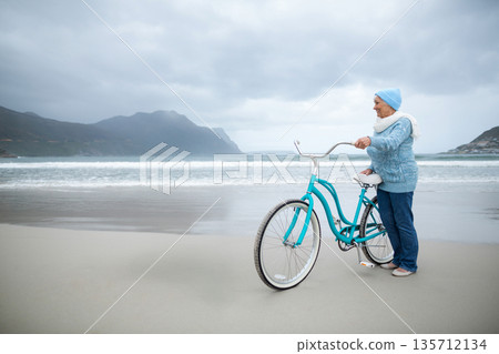 Senior woman holding teal bicycle on beach wearing beanie scarf slip-ons gazing at sea, copy space Senior woman holding teal bicycle on beach wearing beanie scarf slip-ons gazing at sea, copy space 135712134