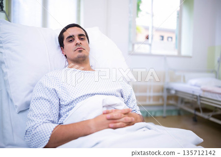Mid adult man reclining on hospital bed wearing patterned gown near window and empty bed Mid adult man reclining on hospital bed wearing patterned gown near window and empty bed 135712142