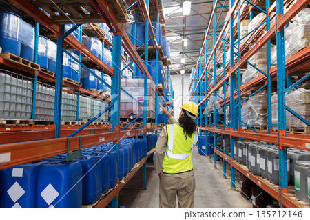 Female warehouse worker wearing safety vest inspecting drums and pallets in warehouse aisle Female warehouse worker wearing safety vest inspecting drums and pallets in warehouse aisle 135712146