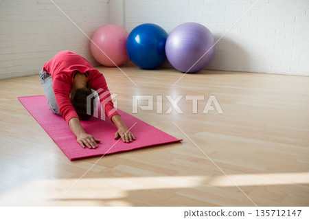Woman wearing red top performing child's pose on pink yoga mat in gym with fitness balls Woman wearing red top performing child's pose on pink yoga mat in gym with fitness balls 135712147