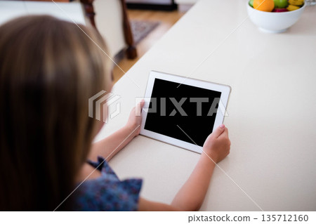 Female child sitting on home kitchen countertop holding white tablet near colorful fruit bowl Female child sitting on home kitchen countertop holding white tablet near colorful fruit bowl 135712160