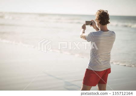 Man in striped shirt, red swim shorts capturing sea horizon on smartphone at beach, copy space Man in striped shirt, red swim shorts capturing sea horizon on smartphone at beach, copy space 135712164