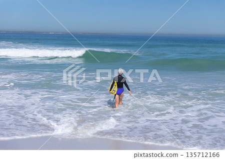 Senior white woman wearing black rashguard and blue bottoms holding yellow bodyboard wading in surf Senior white woman wearing black rashguard and blue bottoms holding yellow bodyboard wading in surf 135712166