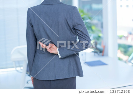 Businesswoman standing at office desk crossing fingers behind back near window blinds and folder 135712168