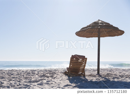 Wooden lounge chair draped red towel standing under straw parasol on beach facing sea, copy space Wooden lounge chair draped red towel standing under straw parasol on beach facing sea, copy space 135712180