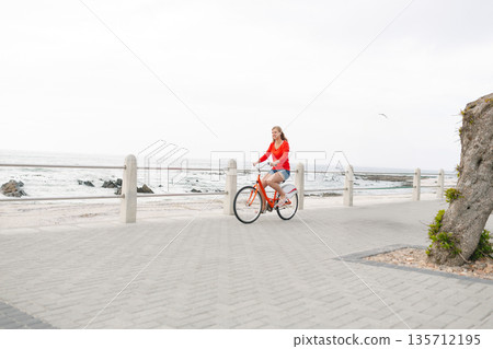 Female cyclist riding orange bicycle along seaside promenade by concrete railing with cloudy sky Female cyclist riding orange bicycle along seaside promenade by concrete railing with cloudy sky 135712195
