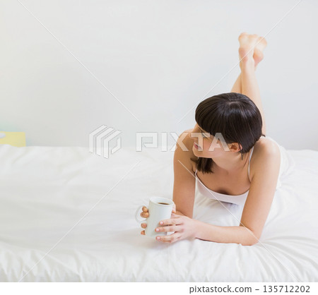 Woman lying on stomach holding white mug while wearing camisole on bed in bedroom, copy space 135712202