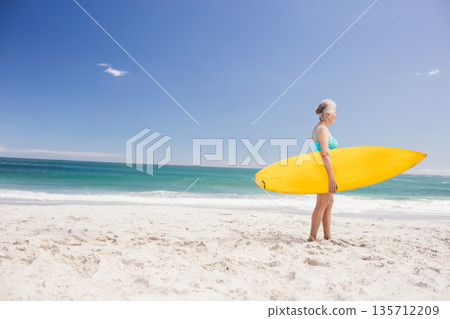 Senior woman holding yellow surfboard in teal swimsuit on beach facing turquoise ocean, copy space Senior woman holding yellow surfboard in teal swimsuit on beach facing turquoise ocean, copy space 135712209
