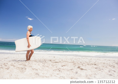 Senior woman standing barefoot on beach holding foam bodyboard facing turquoise ocean, copy space 135712212